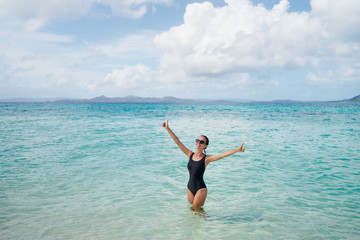 Best vacation! Happy young woman in black swimsuit showing thumb up while swimming on tropical beach.