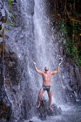 Fototapeta na wymiar Happy young man relaxing under tropical waterfall with arms up raised in freedom. Health and relaxation.