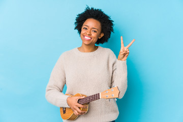 Young african american woman playing ukelele isolated joyful and carefree showing a peace symbol with fingers.