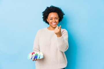 Middle age african american woman eating macaroons isolated pointing with finger at you as if inviting come closer.