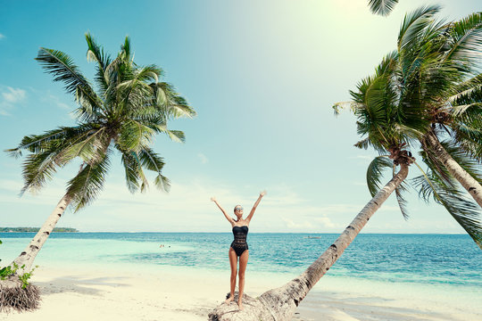 Vacation on the seashore.Young woman in black swimsuit on the beautiful tropical beach standing on the coconut palm tree.