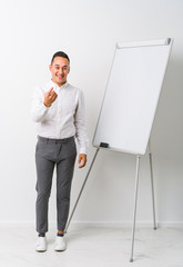 Young latin coaching man with a whiteboard isolated pointing with finger at you as if inviting come closer.