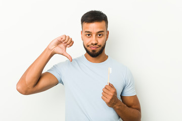 Young south-asian man holding a toothbrush showing a dislike gesture, thumbs down. Disagreement...