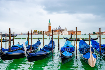 Fototapeta na wymiar Venice, Italy. Gondolas/ Gondole docked by wooden mooring poles. Famous romantic tour boat ride for tourists/ couples/ people. Bell Tower of San Giorgio Maggiore Basilica Church in background.
