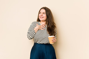 Young curvy woman holding a coffee laughs out loudly keeping hand on chest.