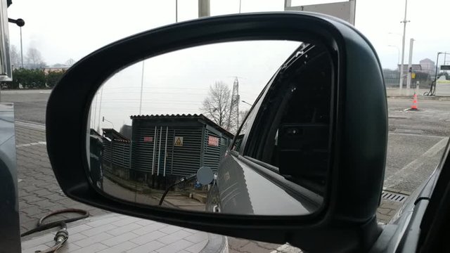 LPG refueling at the distributor: from the rear view mirror you can see the gas pump gun connected to the machine. A car passes the street.