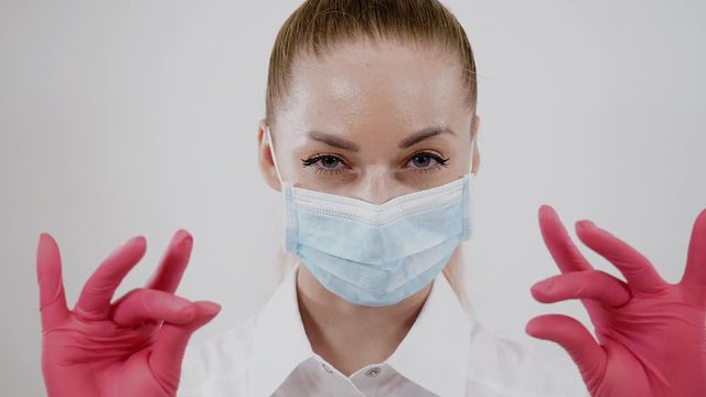 Portrait of young doctor close up isolated on white background. Confident sight by adult masked surgeon and moves gloved fingers. Medical woman raises eyebrow looking at camera. Healthcare concept