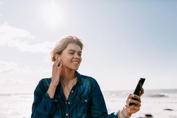 Fascinating smiling girl having fun at beach in weekend morning and posing on sky background with smartphone and listening music