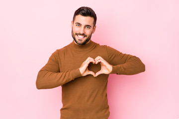 Young caucasian man against a pink background isolated smiling and showing a heart shape with hands.