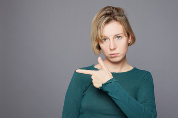 Charming serious woman with thoughtful expression, points aside with index finger, dressed in casual green clothing, has blond hair and blue eyes, isolated over grey background. Look there, please...