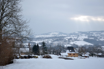 Snowy winter landscape in february 2015. You can see trees, fir trees, woodpile for fireplace, and a field on first plan. There's also a yellow house, with a person training her horse outside. 