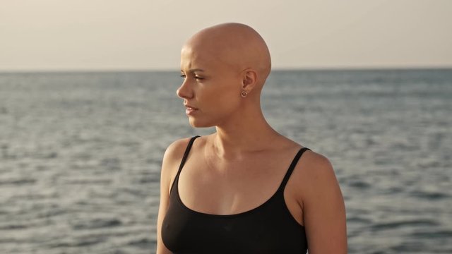Serious attractive bald sports woman looking away while standing near the sea outdoors