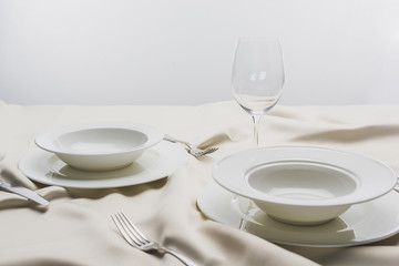 Plates with cutlery and clear wine glass on white tablecloth on grey background