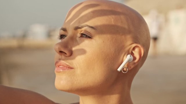 Close up view of Pleased attractive bald sports woman in earphones with finger shadow on the face listening music and looking away while sitting near the sea outdoors