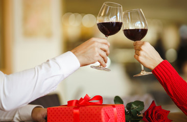 Hands holding glasses of wine on restaurant background. Couple clink glasses with red wine. Valentines day celebration concept. Relationship, surprise and love concept.