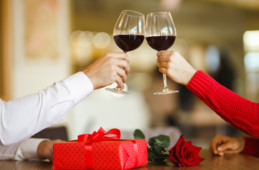 Hands holding glasses of wine on restaurant background. Couple clink glasses with red wine. Valentines day celebration concept. Relationship, surprise and love concept.