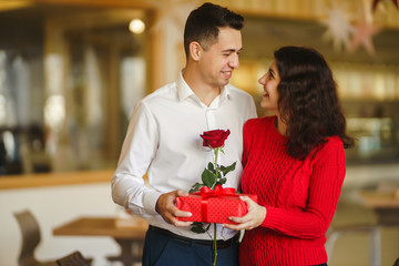 Man gives to his woman a gift box with red ribbon. A loving couple cuddles and celebrating Valentine's Day in the restaurant. Valentine's Day, holiday and surprise concept. Relationship and love.