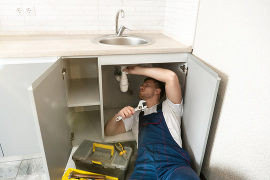 young man plumber in uniform fixing the sink with adjustable spanner in his hand lying on the kitchen floor professional plumbing repair service
