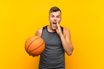 Young handsome blonde man holding a basket ball over isolated yellow background shouting with mouth wide open