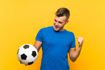 Young handsome blonde man holding a soccer ball over isolated yellow background celebrating a victory