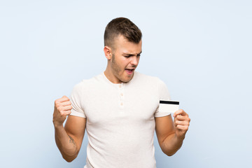 Young handsome blonde man over isolated blue background holding a credit card