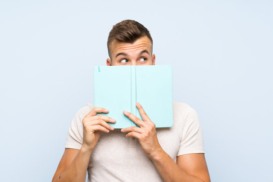 Young handsome blonde man over isolated blue background holding and reading a book