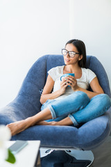 Fototapeta premium Young beautiful woman sitting home in the chair by the window with cup of hot coffee.