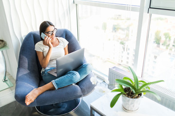 Casual young woman using laptop and cellphone on chair at home