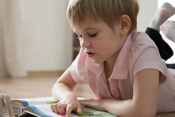 A cute little child, lying on the warm home floor, reading an interesting story in a huge book.
