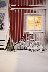 Fototapeta na wymiar Bicycle left under falling snow. Snowy red house with big window and atmospheric lights, Christmas mood photo.