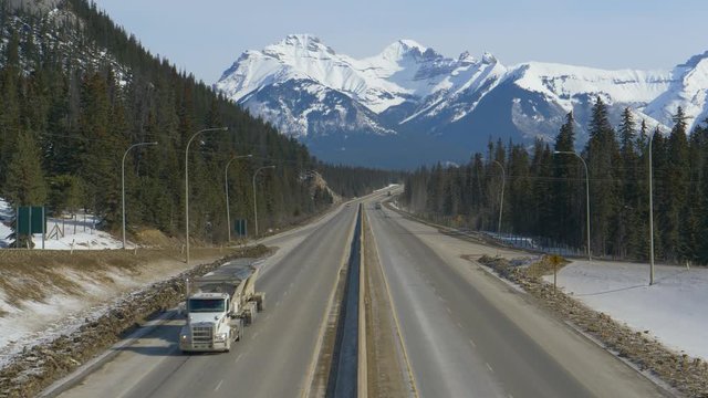 DRONE: Flying above the famous Trans Canada Highway as vehicles drive across the gorgeous wintry landscape. Cars and truck hauling cargo drive away from the stunning snowy mountain range in Alberta.