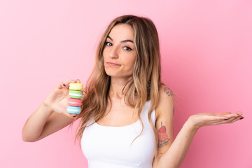Young woman with  macarons over isolated pink background making doubts gesture while lifting the shoulders