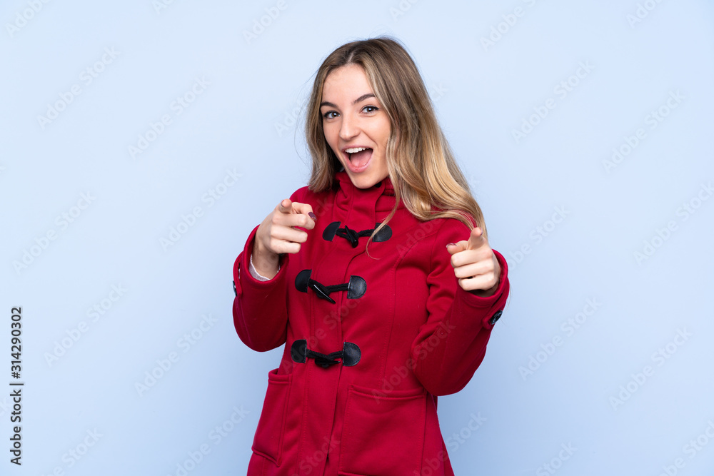 Wall mural young woman with winter coat over isolated blue background points finger at you