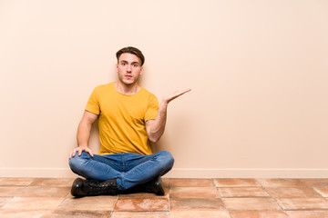 Young caucasian man sitting on the floor isolated impressed holding copy space on palm.