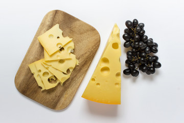 Large peace and many slices of maasdam cheese with large holes  on wooden board and branch of dark blue grapes on white background.