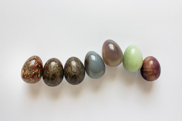 Uneven row of decorative Easter eggs made of precious colored textured stones arranged on white background.