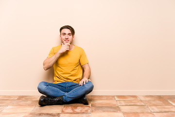 Young caucasian man sitting on the floor isolated smiling happy and confident, touching chin with hand.