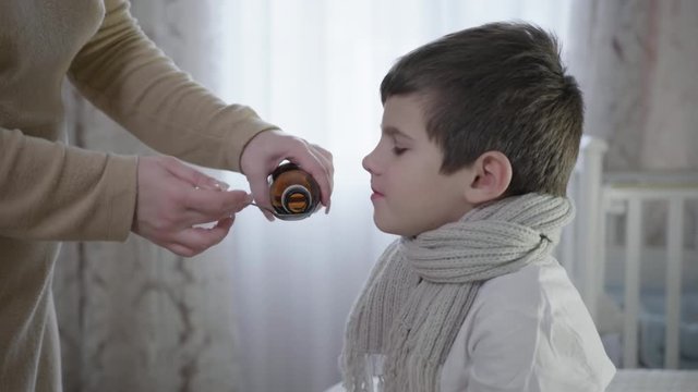 care of children, parent pours cough syrup into a spoon for sick kid takes medicine for the disease sitting on the bed in a warm scarf close up