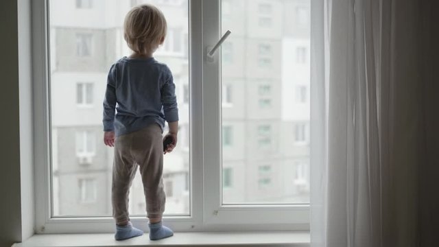 dangerous situation with child at home, little boy stands on the windowsill and looking out the window with toy car in hand