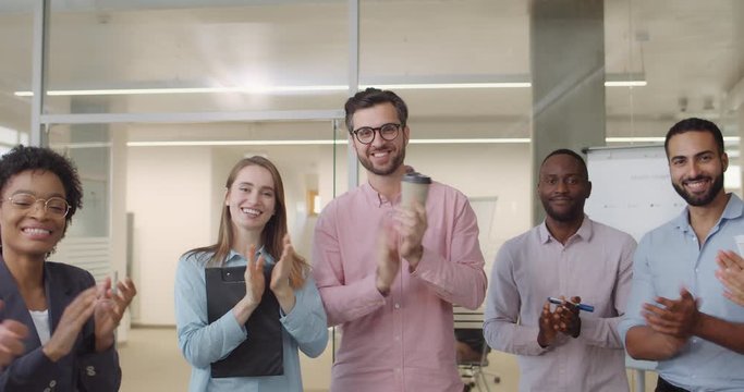 Close up of smiling happy group of diverse workers applouding and standing in office room. Multi ethnic male and female office workers looking happy greeting somebody.