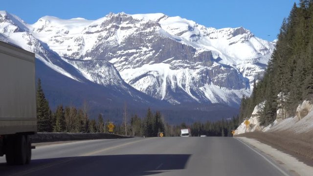 Three trucks haul cargo down the highway leading towards the breathtaking snow capped mountains in Jasper National Park. Lorries travel along the famous Icefields Parkway on a sunny winter day.