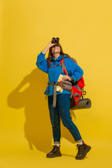 Smiling, looking up. Portrait of a cheerful young caucasian tourist girl with bag and binoculars isolated on yellow studio background. Preparing for traveling. Resort, human emotions, vacation.