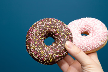 Hand holding two a delicious colorful donut with sprinkles near dark blue background, sugar concept pink and brown