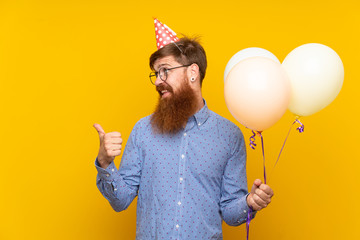 Redhead man with long beard holding balloons over isolated yellow background pointing to the side to present a product