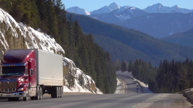 Red freight truck hauls a container across the beautiful Jasper National Park on a sunny spring afternoon. Big rig transports cargo across the pine forest covering the valley under snowy mountains.