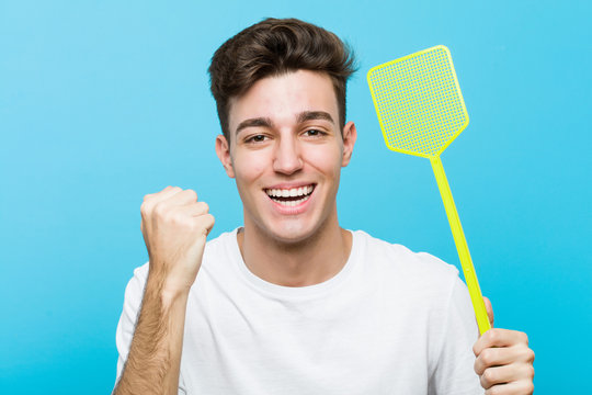 Young caucasian man holding a fly swatter cheering carefree and excited. Victory concept.