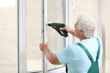 Mature construction worker repairing plastic window with electric screwdriver indoors