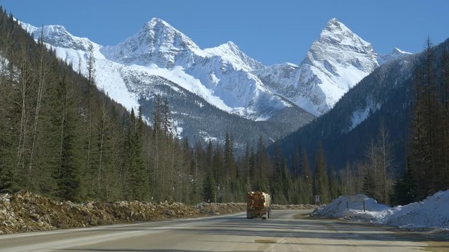Cars and truck hauling logs drives along road running under snowy mountain range in Alberta. Spectacular snow covered mountains tower above a scenic road leading across the rugged Canadian wilderness.