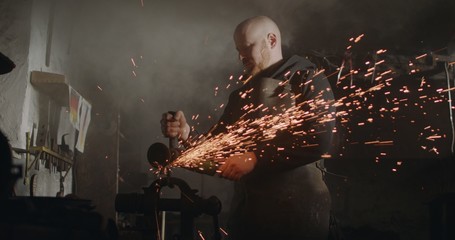 Young blacksmith with angle grinder sparks in his workshop dark eerie haze