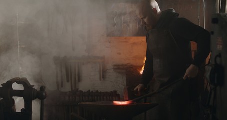 Blacksmith hammering heated iron on anvil making knife dark eerie haze